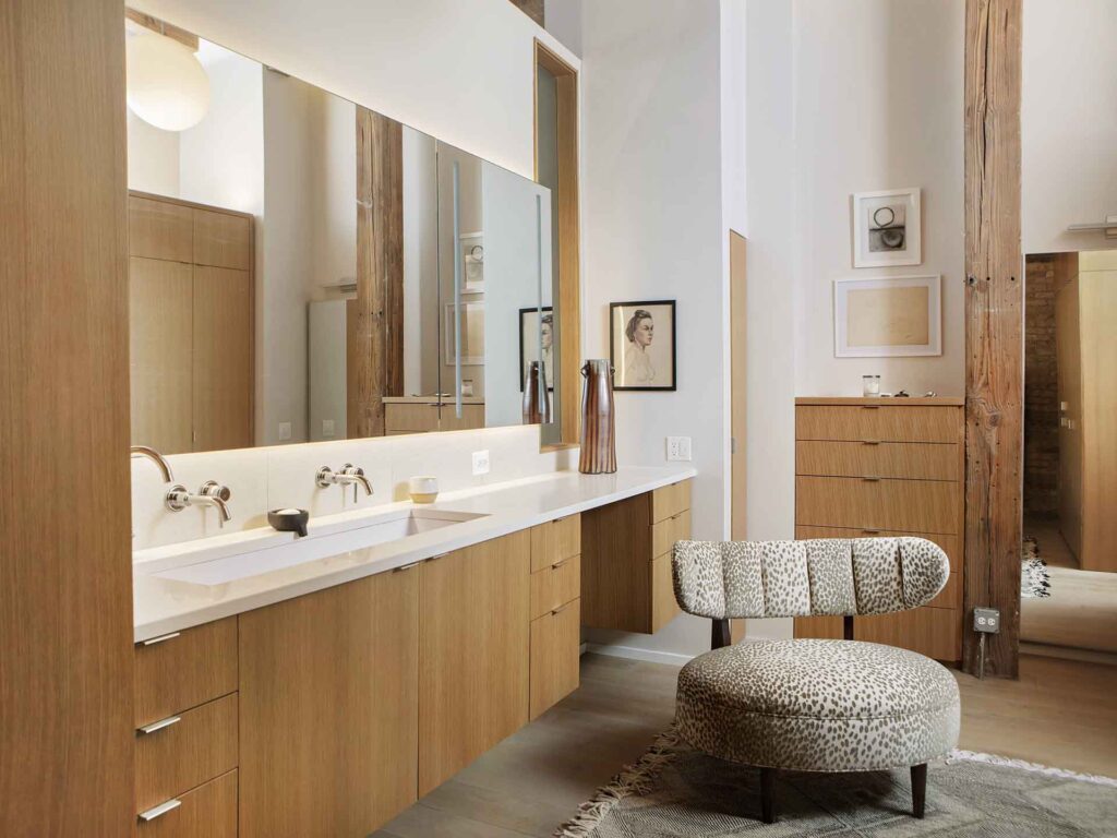A modern bathroom with light wood cabinets, a large mirror, wall-mounted sinks, and a patterned chair. Minimalist décor includes framed art on white walls and exposed wooden beams.