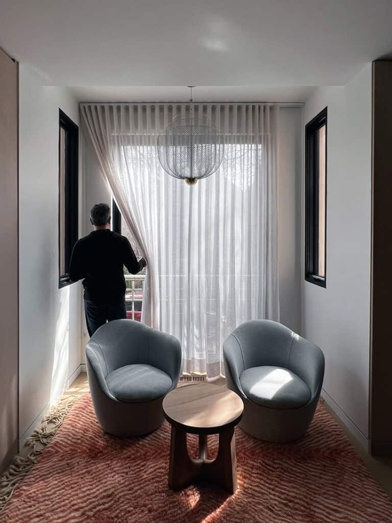 A person stands by sheer curtains in a bright room with two gray armchairs, a small wooden table, and orange patterned carpet.