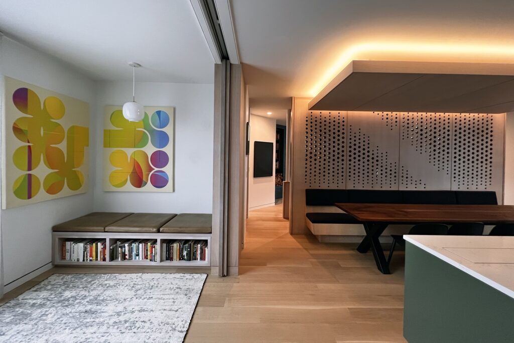 A modern living space with abstract colorful art on white walls, a built-in bench with cushions and books underneath, a light wood floor, and a dining area with a walnut wooden table and geometric perforated wall panels.