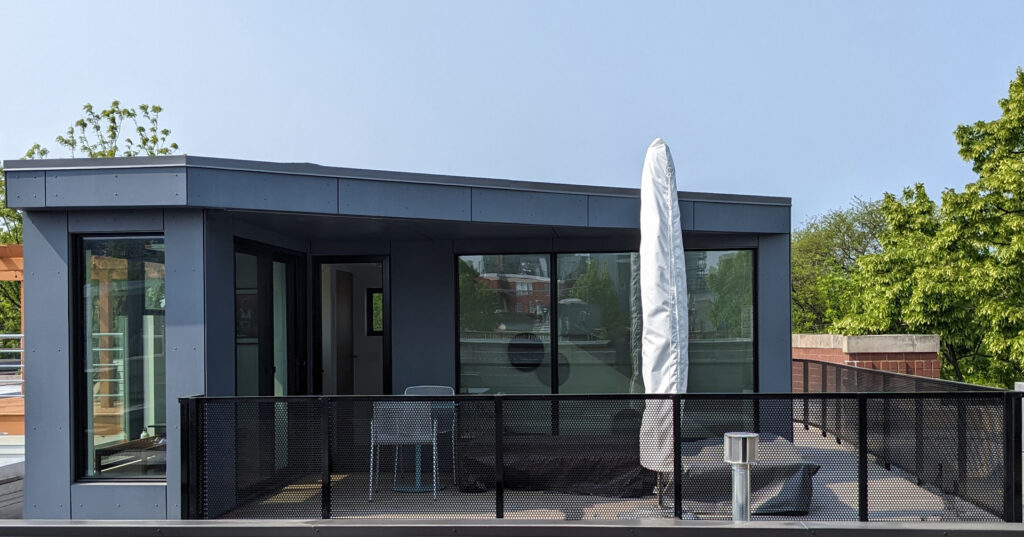 Modern rooftop deck with a small gray structure featuring large windows and doors, a covered umbrella, two chairs, and metal railing; trees and blue sky in the background.