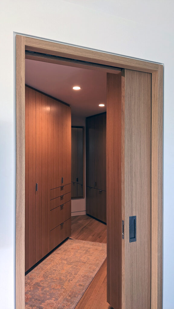 A view through a partially open wooden sliding pocket door reveals a modern walk-in closet with built-in wooden cabinets, drawers, a mirror, and a light-colored patterned rug on the floor.
