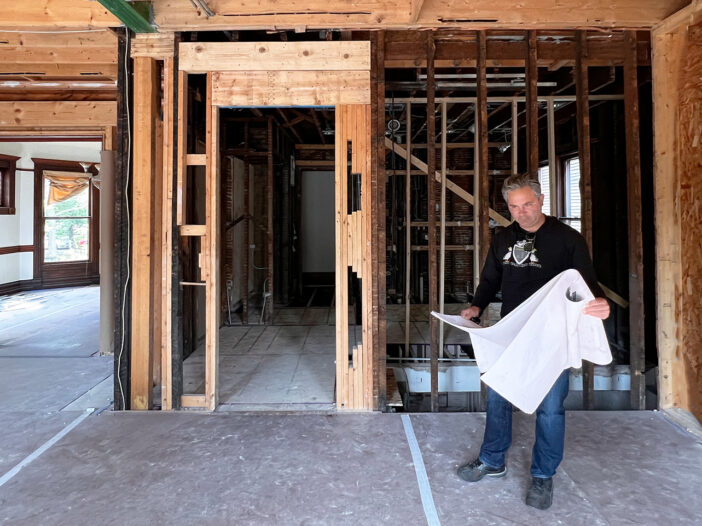 A male architect stands in a partially constructed room holding blueprints, surrounded by exposed wooden framing and beams. The image has the moss logo in the bottom right corner.