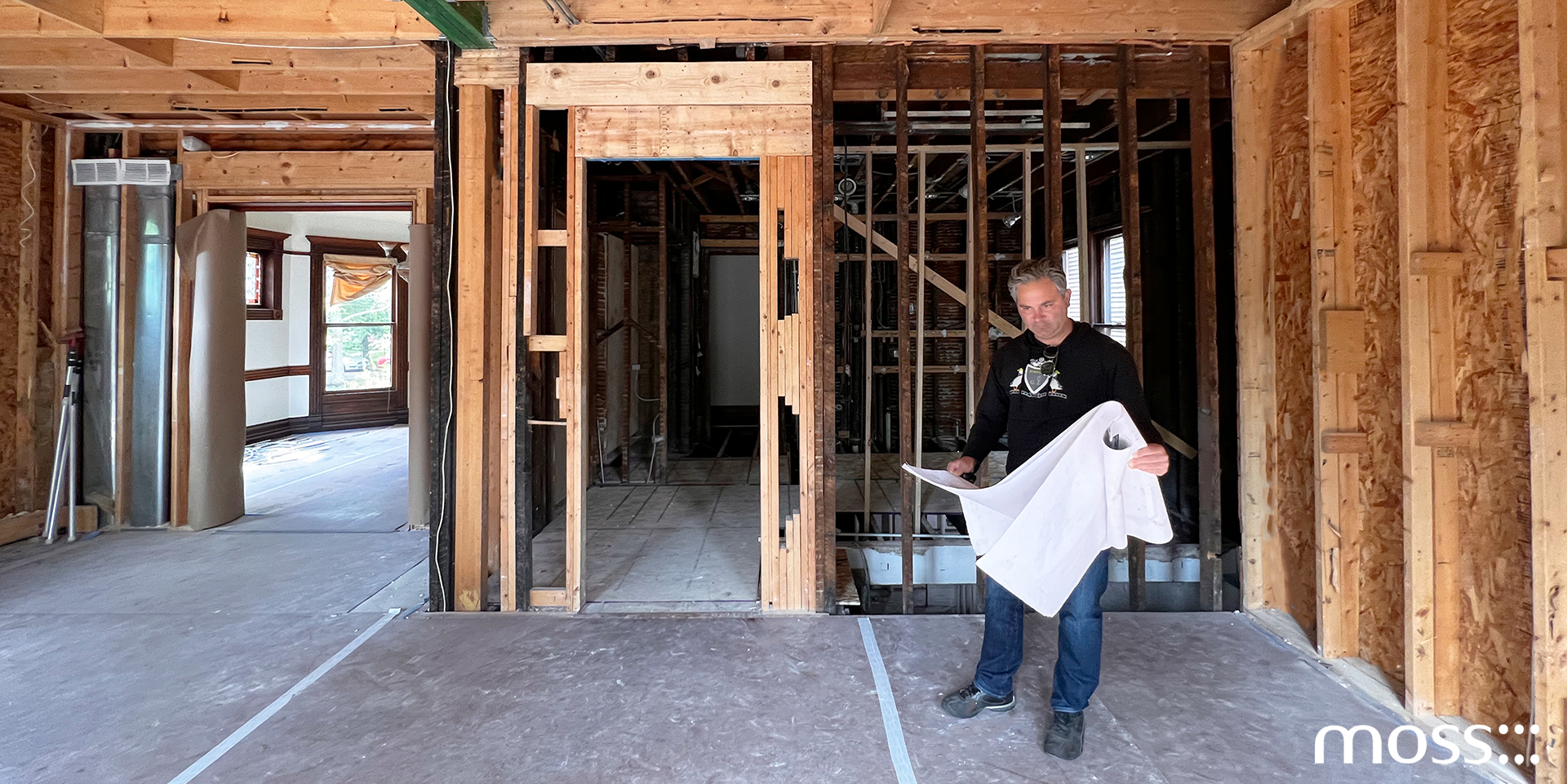 A male architect stands in a partially constructed room holding blueprints, surrounded by exposed wooden framing and beams. The image has the moss logo in the bottom right corner.