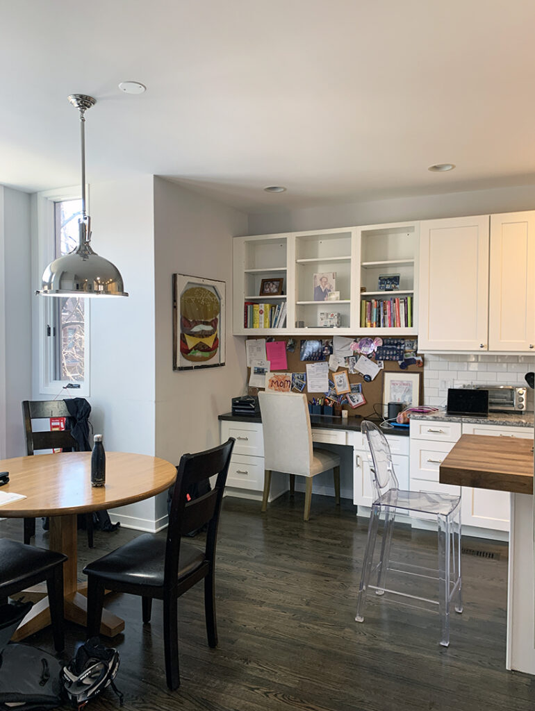 A round wooden table with four dark chairs sits in a bright dining area in a kitchen with large windows, white cabinets, a hanging metal lamp above, and backpacks on the floor near the chairs.