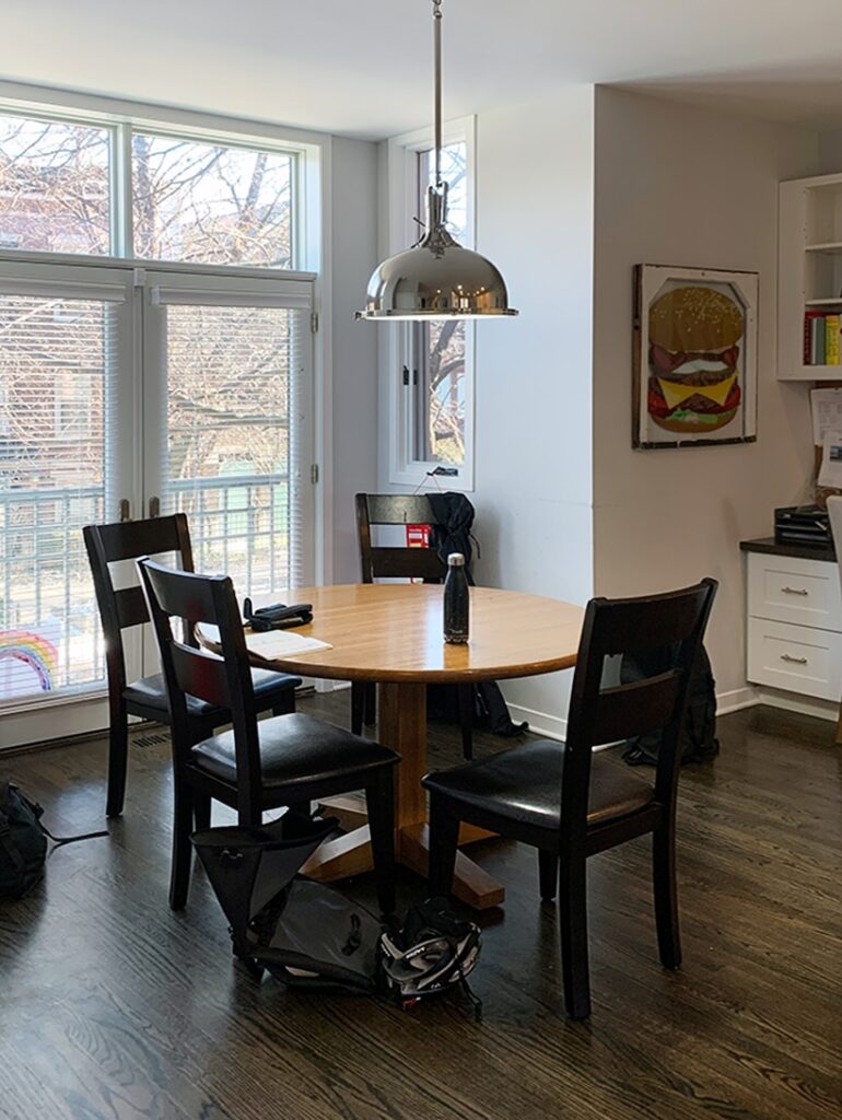 A round wooden dining table with four black chairs sits in a bright room with large windows. A silver pendant light hangs above, and bags are on the floor around the table. Art and shelves are visible in the background.