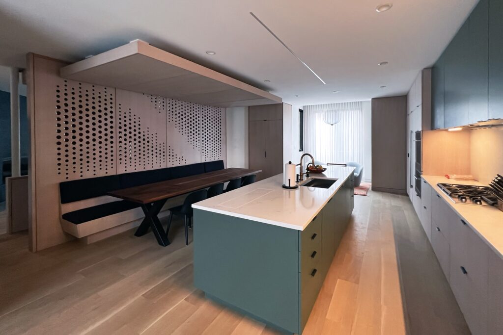Modern kitchen and dining area with a marble island, fruit bowl, built-in sink, and stove. Wooden cabinets, long dark walnut dining table with black chairs are visible under warm, recessed lighting with a wood perforated dividing all behind the dinng table.