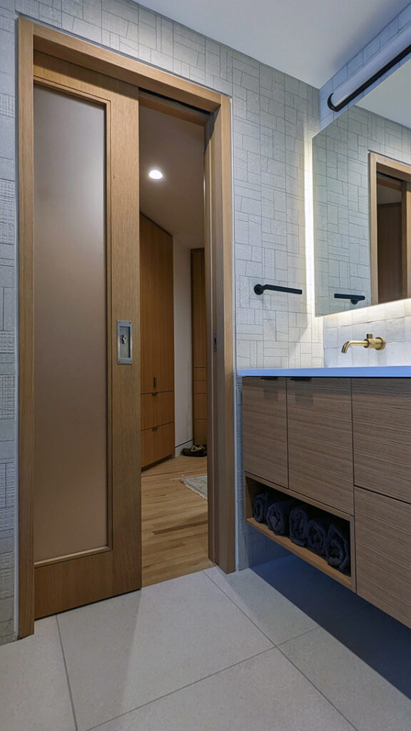A modern bathroom with a wood vanity, white oak sliding pocket door, view to walk in closet area, and a tiled wall.