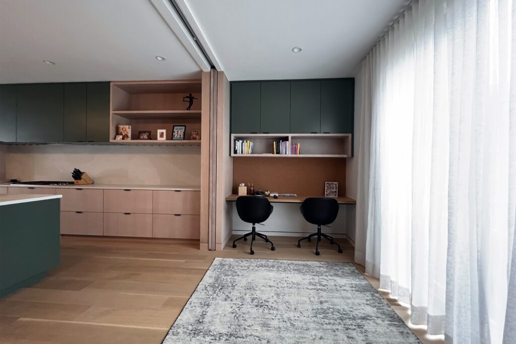 Modern kitchen and worktop computer area separated by a concealed wooden pocket door. Kitchen island with white marble counters, a mix of green and light wood cabinets, with black chairs tucked under working desk, mixed gray area rug and white linen curtains.