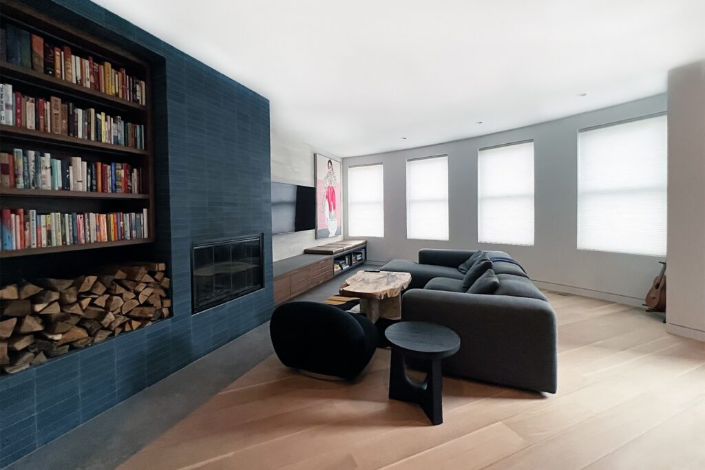 An open, modern living room with light wood floor, dark couch, windows and a blue tiled wall that has a book case with books and a built in fireplace next to logs for the fireplace.