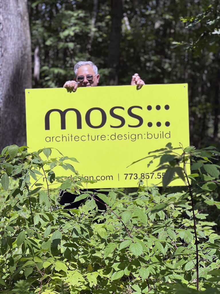 A person wearing sunglasses holds a bright yellow sign reading moss: architecture: design: build while standing among green plants in a wooded outdoor setting.