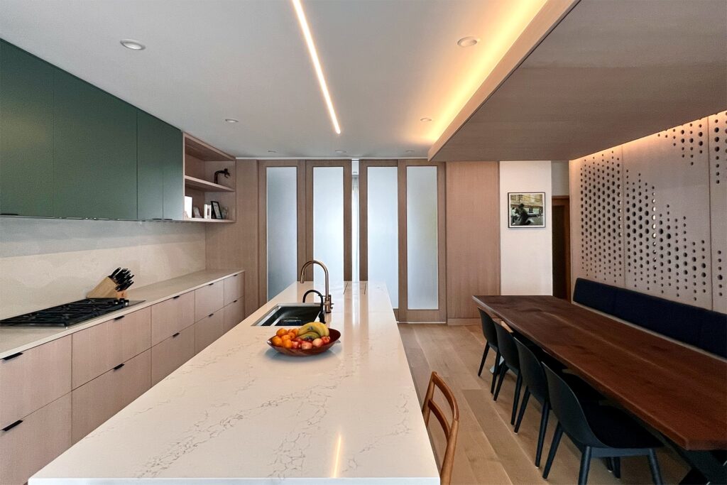 Modern kitchen and dining area with a marble island, fruit bowl, built-in sink, and stove. Wooden cabinets, frosted glass siding doors, and a long dark walnut dining table with black chairs are visible under warm, recessed lighting