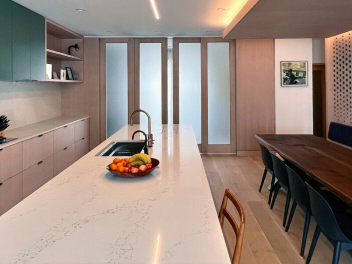 Modern Townhome Renovation with a modern kitchen and dining area with a marble island, fruit bowl, built-in sink, and stove. Wooden cabinets, frosted glass siding doors, and a long dark walnut dining table with black chairs are visible under warm, recessed lighting