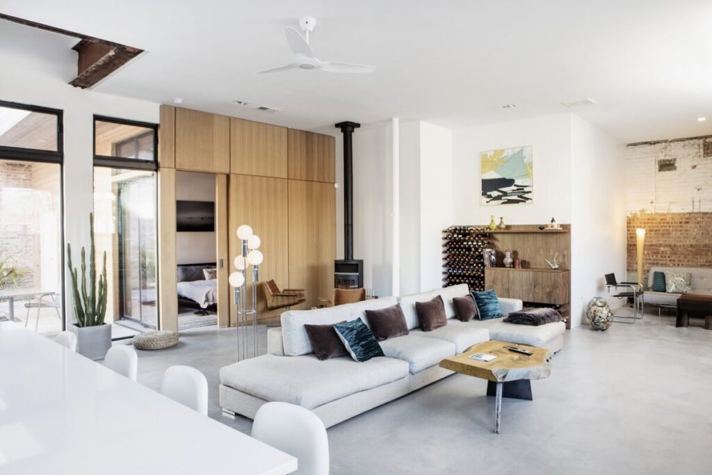 Moss Chicago Architect Architect In Chicago Vs Architect Near Me. Modern living room with a spacious white sofa adorned with colorful pillows, a wooden coffee table, and minimalist decor. A fireplace stands against the wall, while large windows bring in natural light. The room exudes warmth and comfort with its stylish, contemporary design.